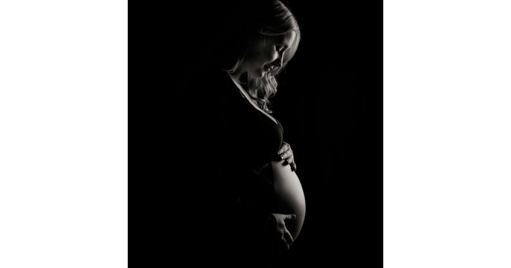 Na imagem há uma mulher grávida segurando a sua barriga, a imagem é preto e branco. A imagem está representando " como a gestação impacta na saúde capilar"